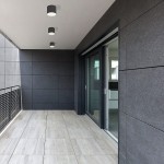 Hexa lampa elewacyjna ogrodowa 1-punktowa w kolorze grafitowym, umieszczona na ścianie, okrągła, z gładką powierzchnią. W tle widoczna szklana ściana oraz płytki na podłodze. Lampa w nowoczesnym stylu, idealna do aranżacji przestrzeni zewnętrznych.