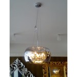 Lampa wisząca Crystal, 5-punktowa, w kolorze chromu, z dużym, okrągłym kloszem. Zawieszona na cienkich linkach, widoczna elegancka struktura wnętrza. Tło w jasnym kolorze, w otoczeniu dekoracyjnego lustra i złotych elementów.