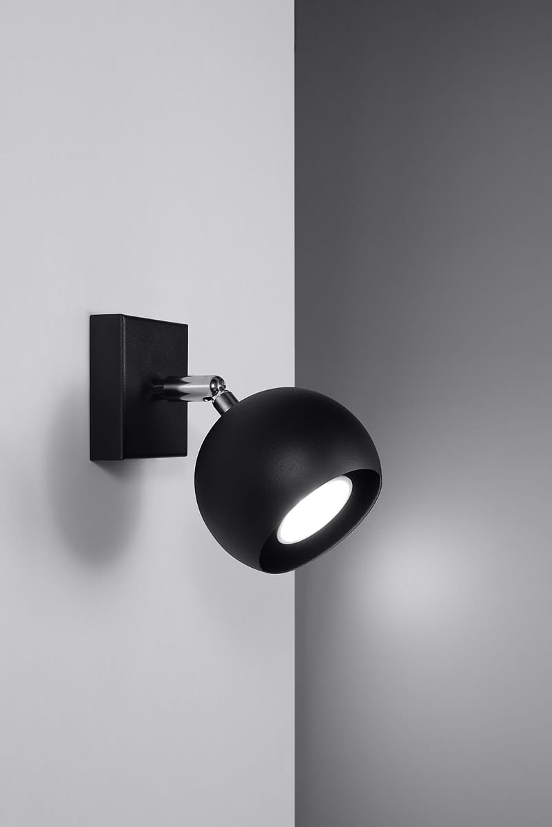 Kinkiet Oculare w kolorze czarnym z okrągłą głowicą lampy. Ustawiony frontalnie na ścianie, z metalowym mocowaniem i chromowanym przegubem. Wydobywa światło z wnętrza, tworząc minimalistyczny efekt. Tło w odcieniach szarości.