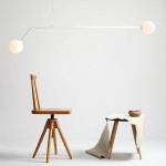 Biała lampa wisząca 2-punktowa Pure 1064H z dwoma okrągłymi kloszami. Lampa zawieszona na cienkim, prostym ramieniu, umieszczona nad drewnianym stołkiem i krzesłem. Tło jasne, minimalistyczne, podkreślające nowoczesny design.