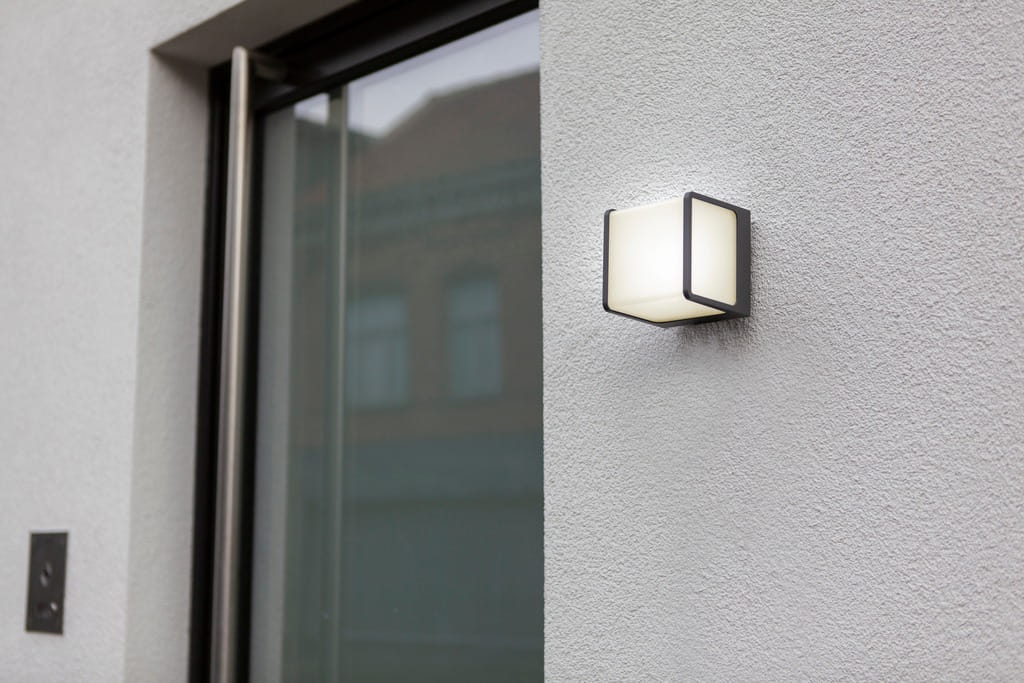 Kinkiet elewacyjny Doblo LED 3000K w kolorze ciemnoszarym, zamontowany na ścianie. Posiada prostokątną, białą osłonę z matowego materiału, z widocznym metalowym ramieniem. Produkt umieszczony obok okna w otoczeniu gładkiej powierzchni ściany.