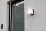 Kinkiet elewacyjny Doblo LED 3000K w kolorze ciemnoszarym, zamontowany na ścianie. Posiada prostokątną, białą osłonę z matowego materiału, z widocznym metalowym ramieniem. Produkt umieszczony obok okna w otoczeniu gładkiej powierzchni ściany.