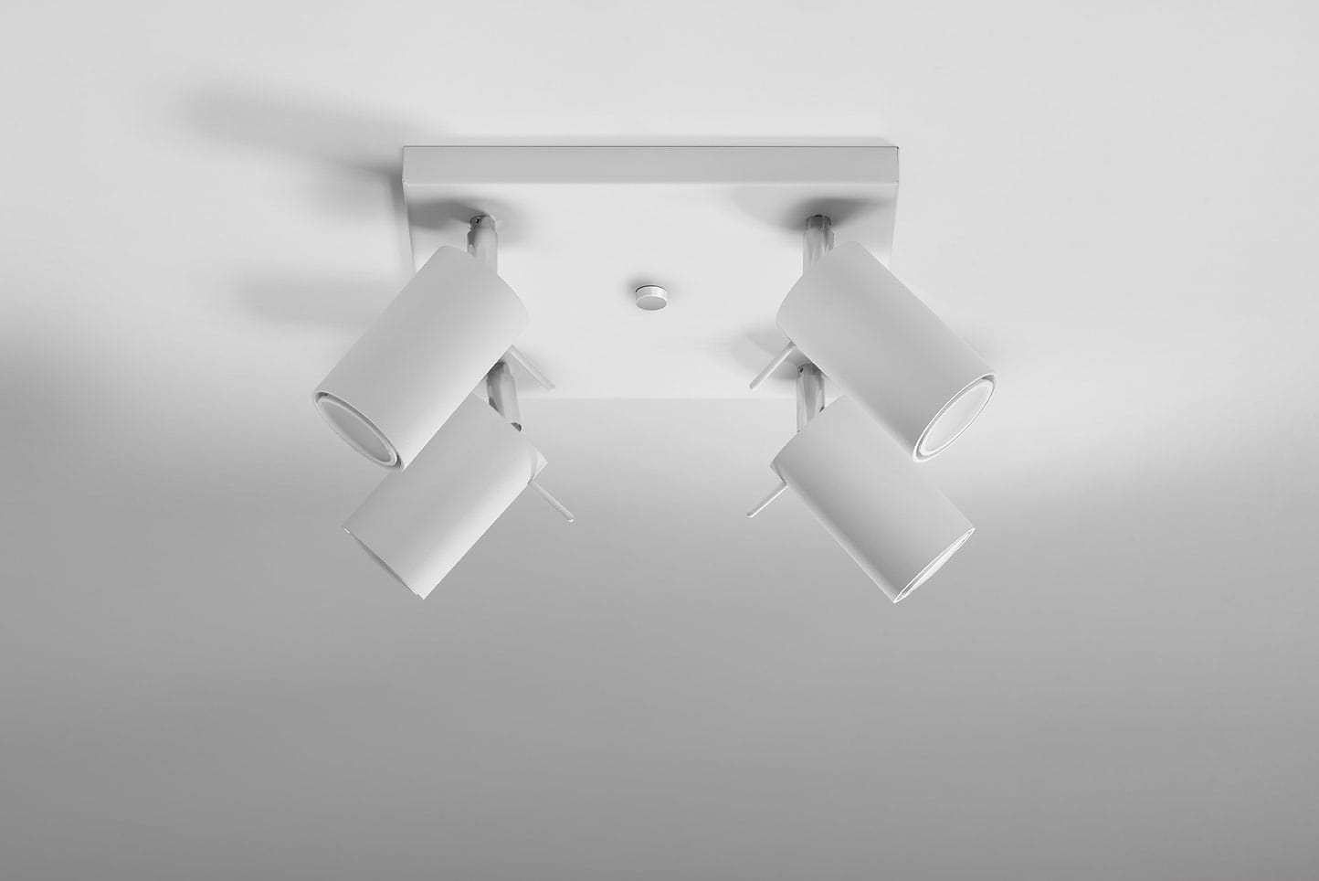 Czteropunktowa lampa sufitowa w kolorze białym z prostokątnym stelażem. Ułożona poziomo, z czterema regulowanymi reflektorami w kształcie cylindrów. Tło gładkie, jasne, co podkreśla design lampy. Idealna do oświetlenia wnętrz.