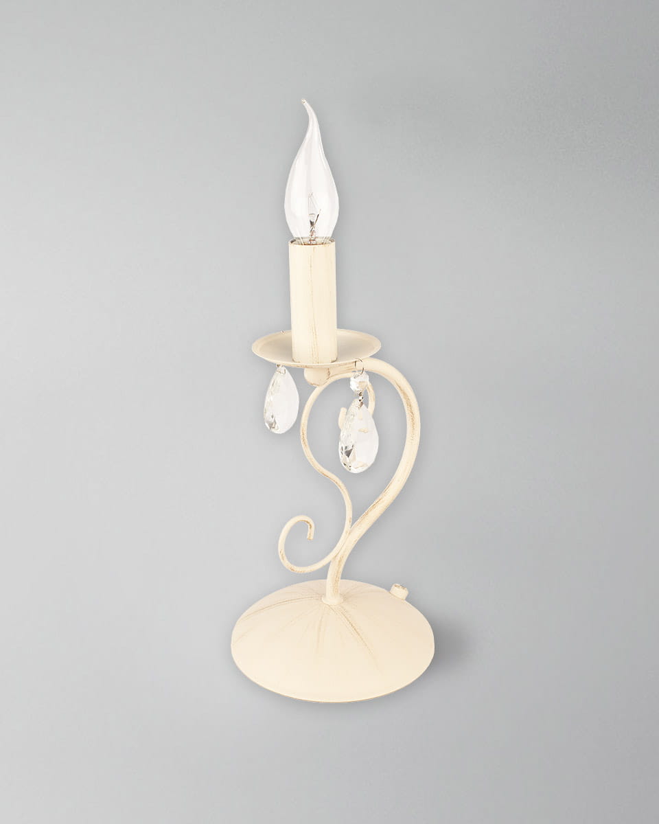 Lampa stołowa Velio 1-punktowa w kolorze kremowym, z metalową podstawą w kształcie okręgu. Wyposażona w przezroczystą żarówkę w kształcie płomienia oraz dwa kryształowe zwisy. Ujęcie z boku, na jednolitym tle.