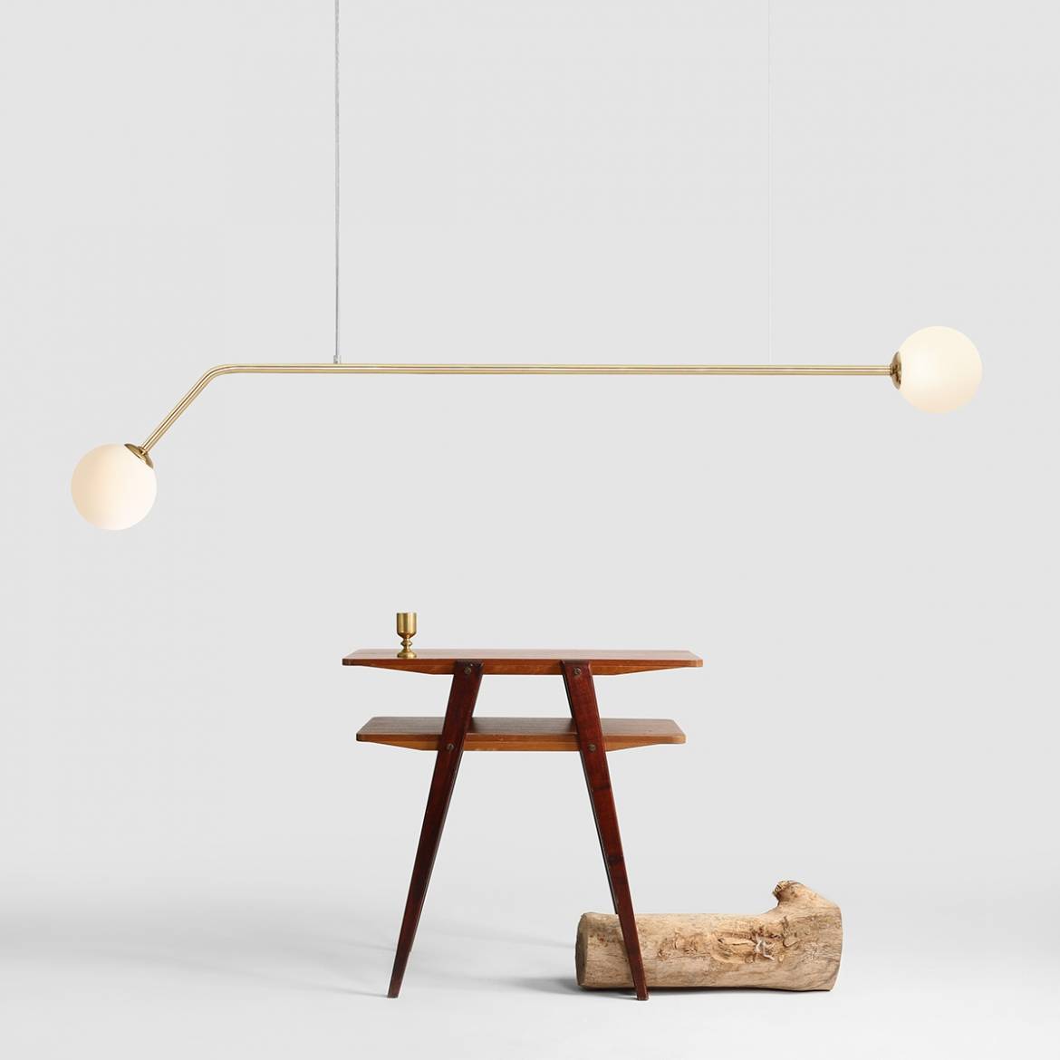 Lampa wisząca 2-punktowa Pure w złotym kolorze, z długim, prostokątnym ramieniem i dwoma okrągłymi kloszami z białego szkła. Produkt zawieszony na cienkim kablu, w tle drewniany stół i kawałek drewna. Tło jasnoszare.