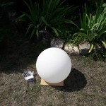 Lampa ogrodowa Cumulus M w kształcie białej, matowej kuli, umieszczona na kartonowym podstawie, z widocznym przewodem elektrycznym obok. Tło z trawnikiem i roślinnością.