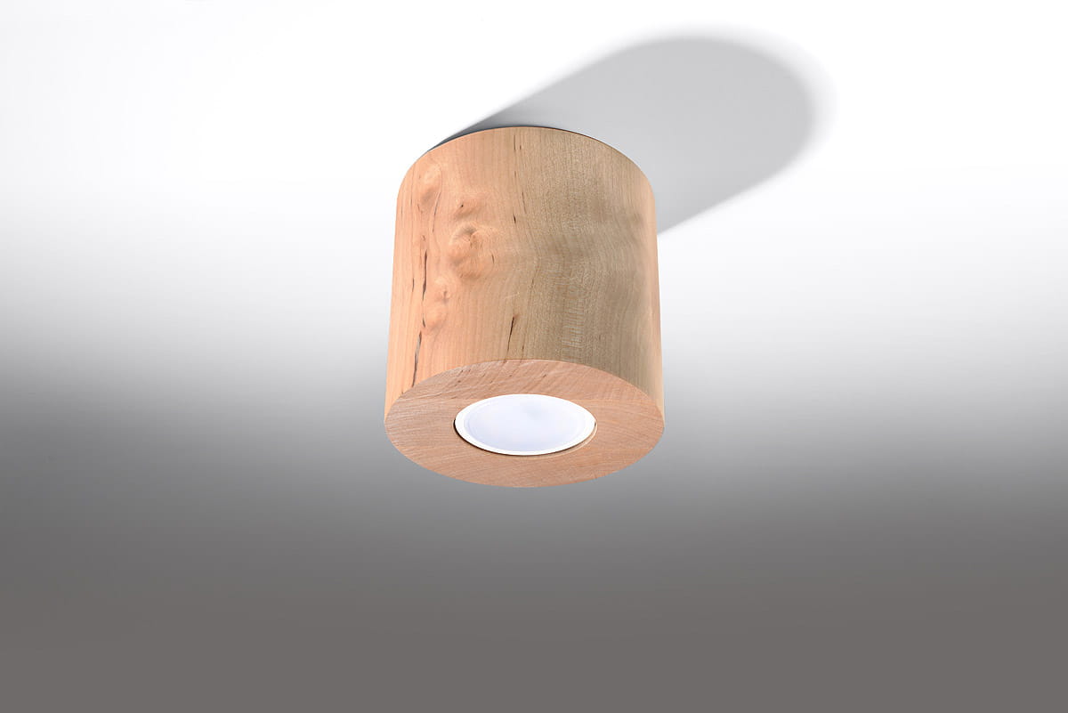 Lampa sufitowa Orbis Drewno 1-punktowa SL.0492 w kształcie cylindrycznym, wykonana z jasnego drewna. Wyposażona w okrągły otwór na źródło światła, zamontowana pod kątem, rzucająca cień na ścianę. Tło w odcieniach szarości.